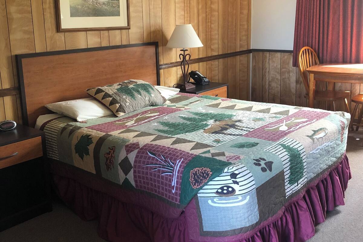 A bedroom with a wooden paneled wall, a bed with a colorful quilt, a nightstand with a lamp, and a chair by a window with a red curtain.