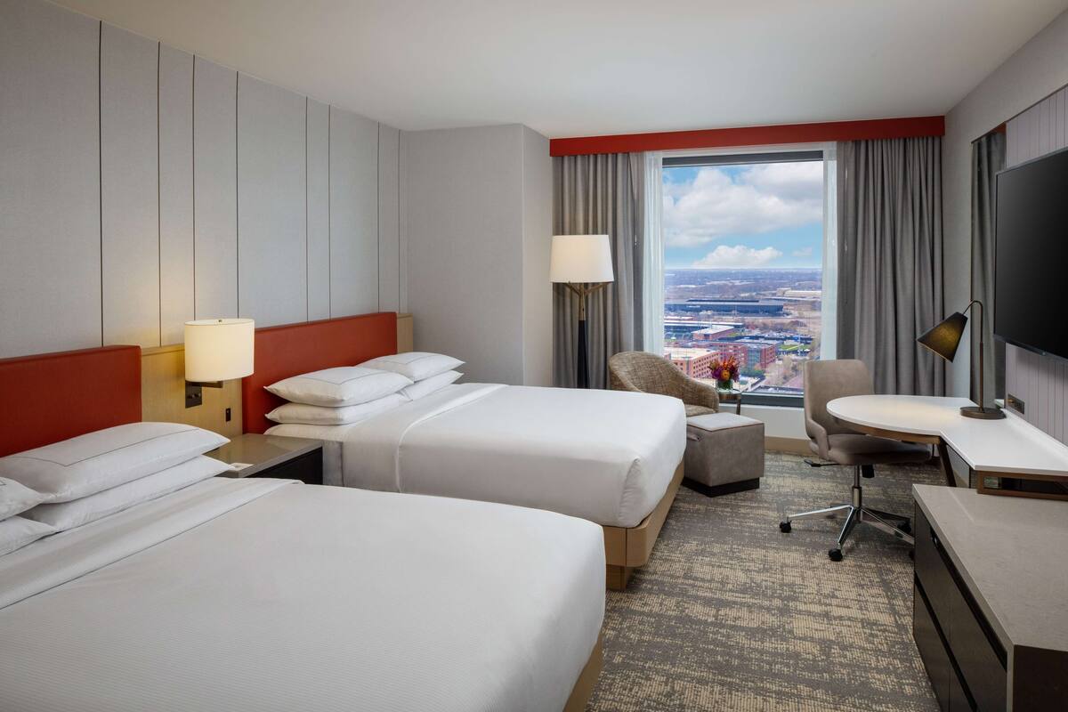 A hotel room with two beds, a desk, a chair, a TV, and a view of the city.