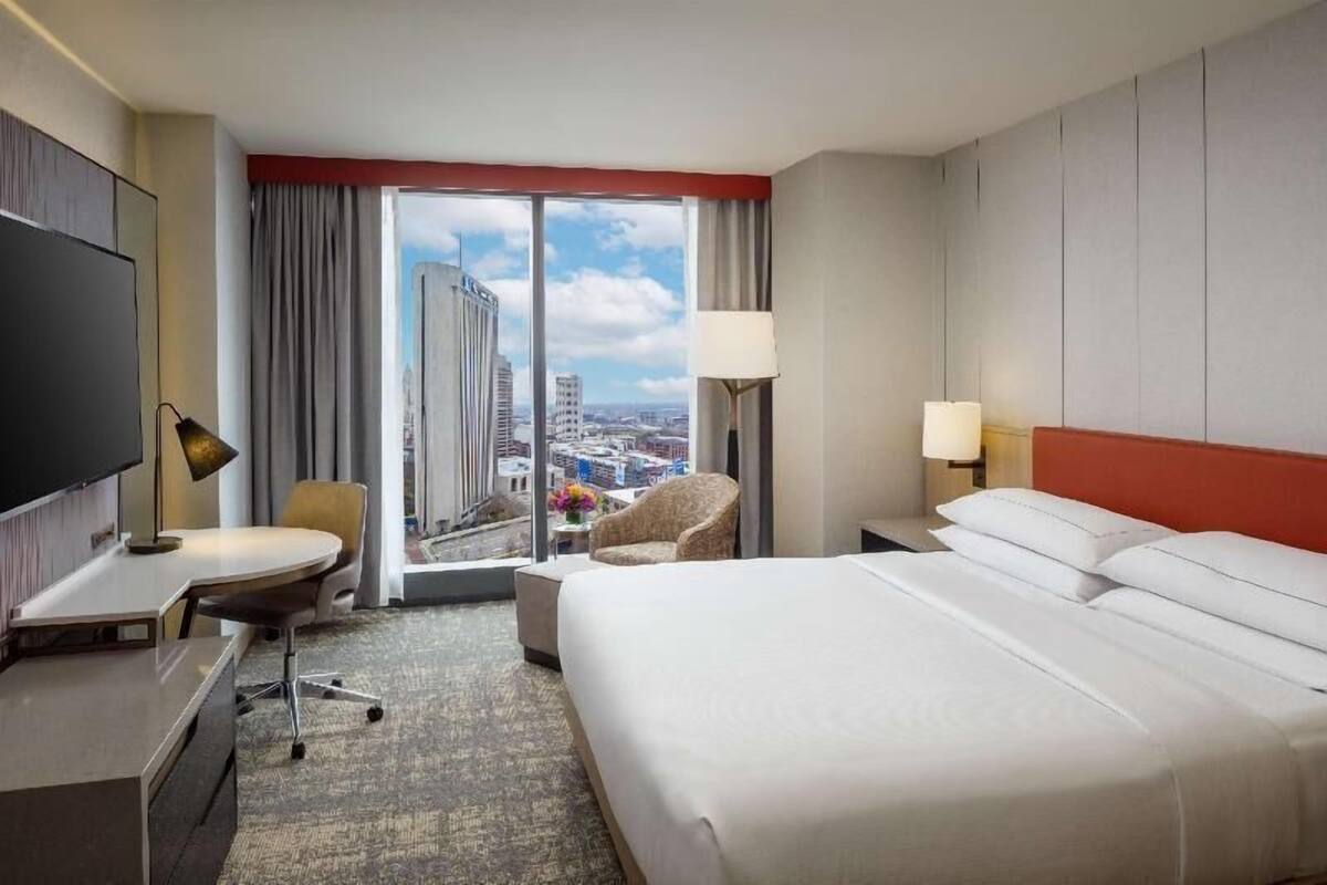 A hotel room with a large bed, a desk, a chair, a TV, and a view of the cityscape.