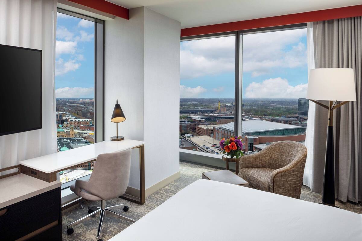 A hotel room with a large window offering a city view, a desk with a chair, a bed, and a decorative vase with flowers.