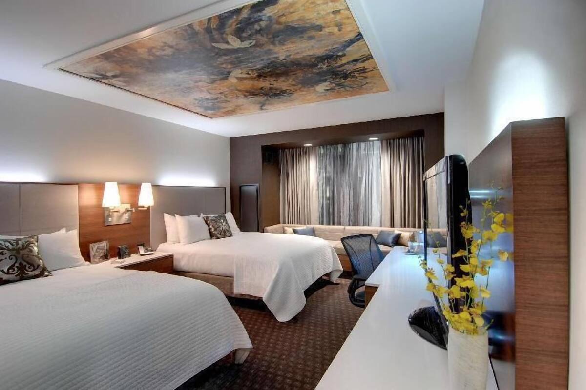 A hotel room with two beds, a desk, a chair, a television, and a painting on the ceiling.
