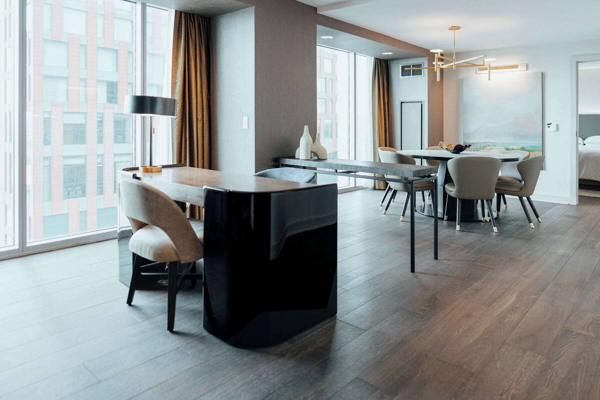 A modern hotel room with a dining area, large windows, and a view of the cityscape.