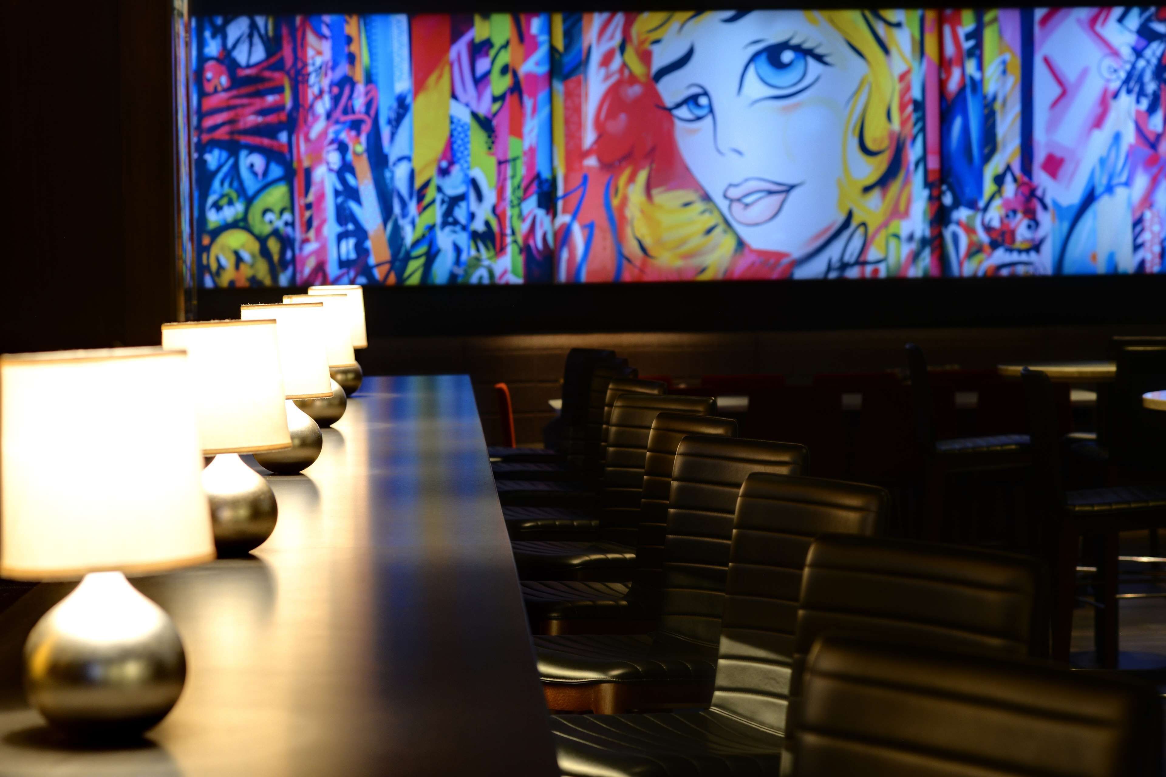 A bar area with a long counter, multiple illuminated lamps, and a large screen displaying a colorful abstract painting.