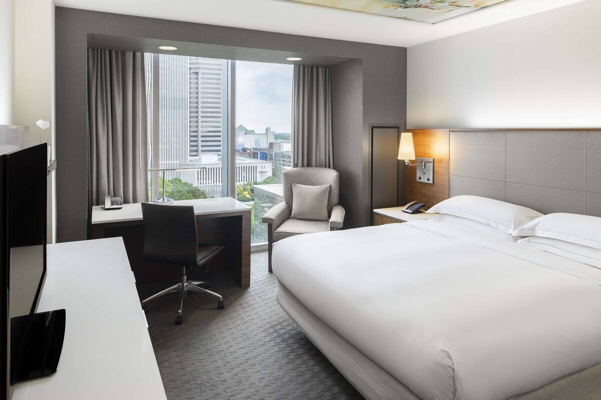 A hotel room with a large bed, a desk with a chair, a lamp, and a view of the city through the window.