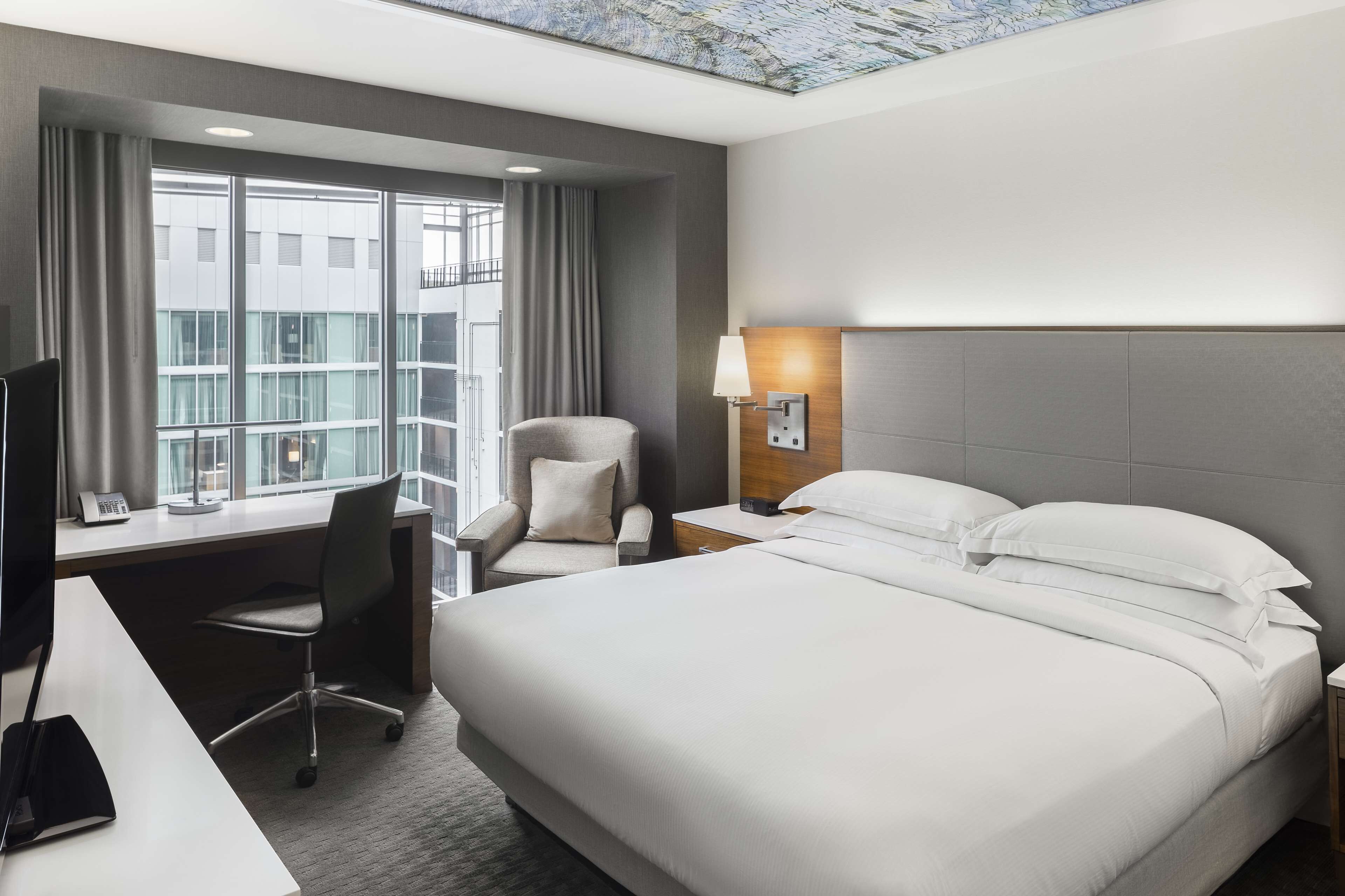 Quarto de hotel com uma cama grande, uma escrivaninha com cadeira, uma luminária e vista da cidade através de amplas janelas.