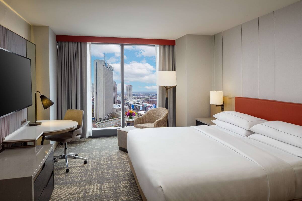 A hotel room with a large bed, a desk, a chair, a TV, and a view of the cityscape.