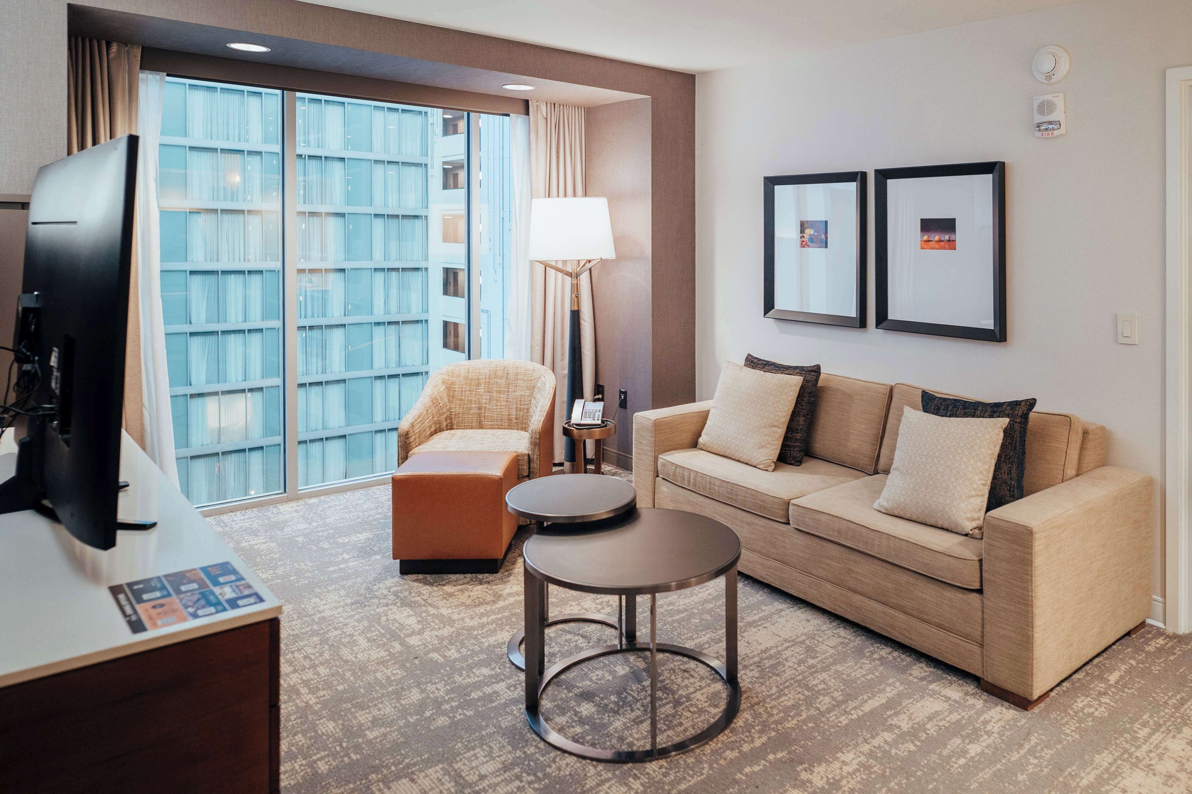 A hotel room with a large window, a sofa, a chair, a small table, and a television.