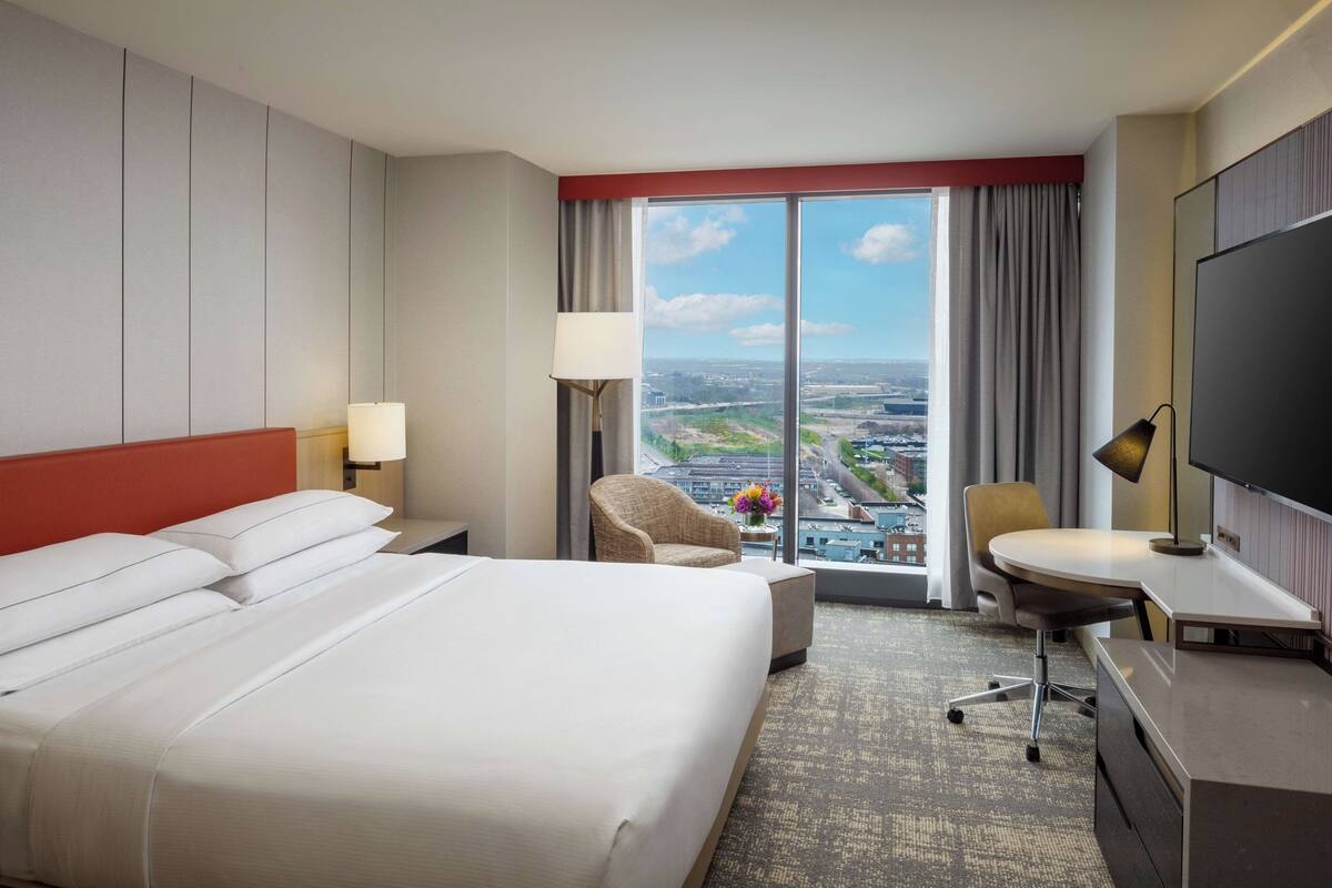 A hotel room with a large bed, a desk, a chair, a TV, and a view of the city through large windows.