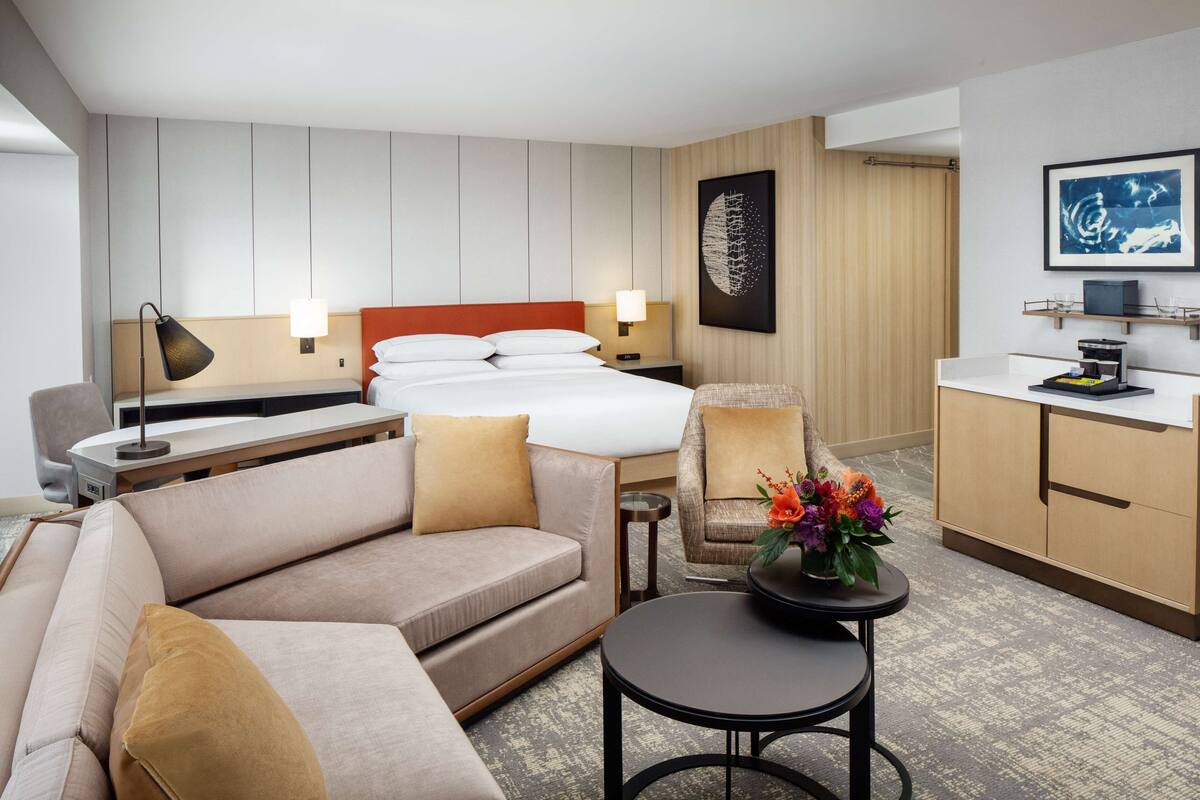 A hotel room with a bed, a sofa, a small table with a vase of flowers, a desk with a lamp, a chair, and a kitchenette area.