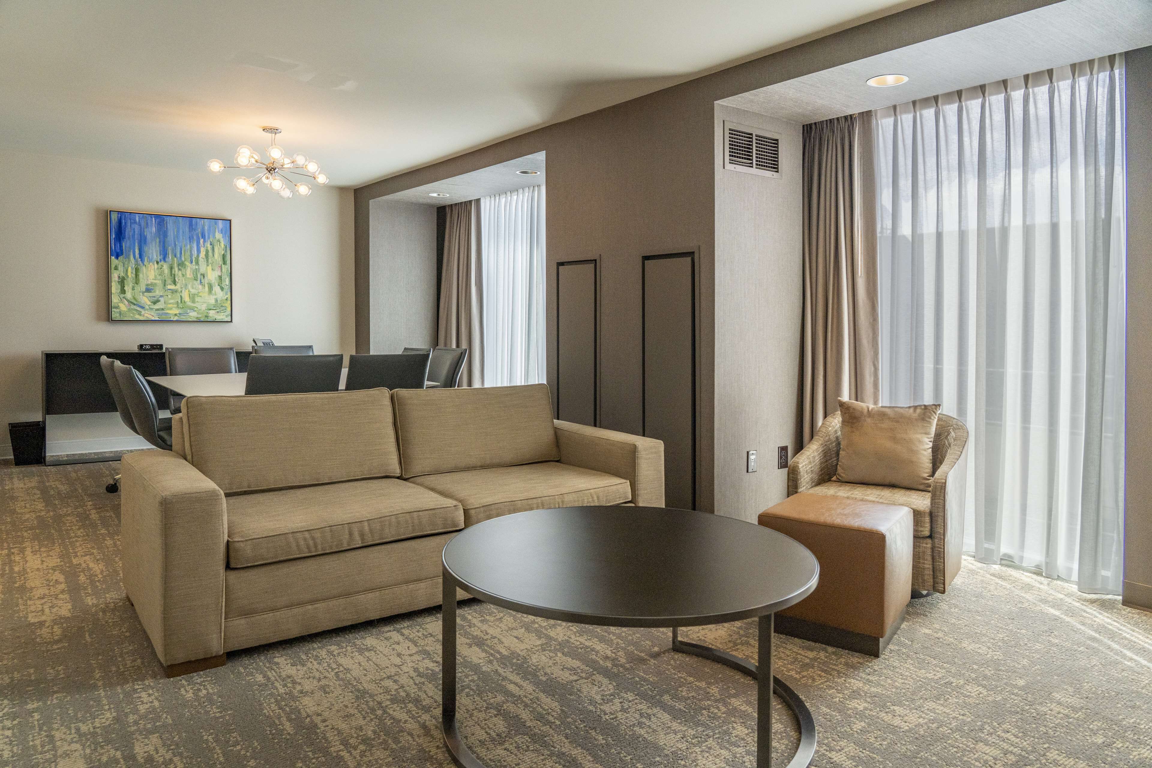 A modern hotel room with a sofa, armchair, and a small table. There is a painting on the wall and a large window with curtains.