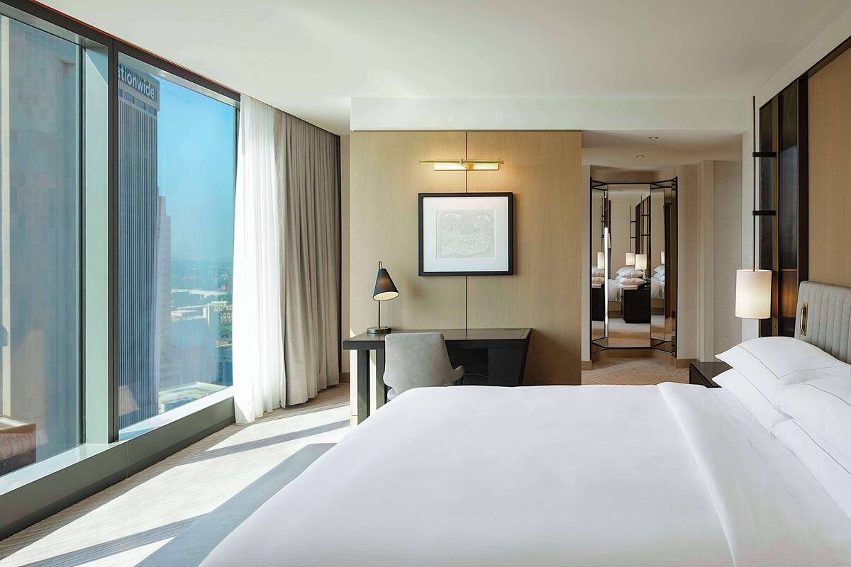 A hotel room with a large bed, a desk, a chair, and a large window with curtains.