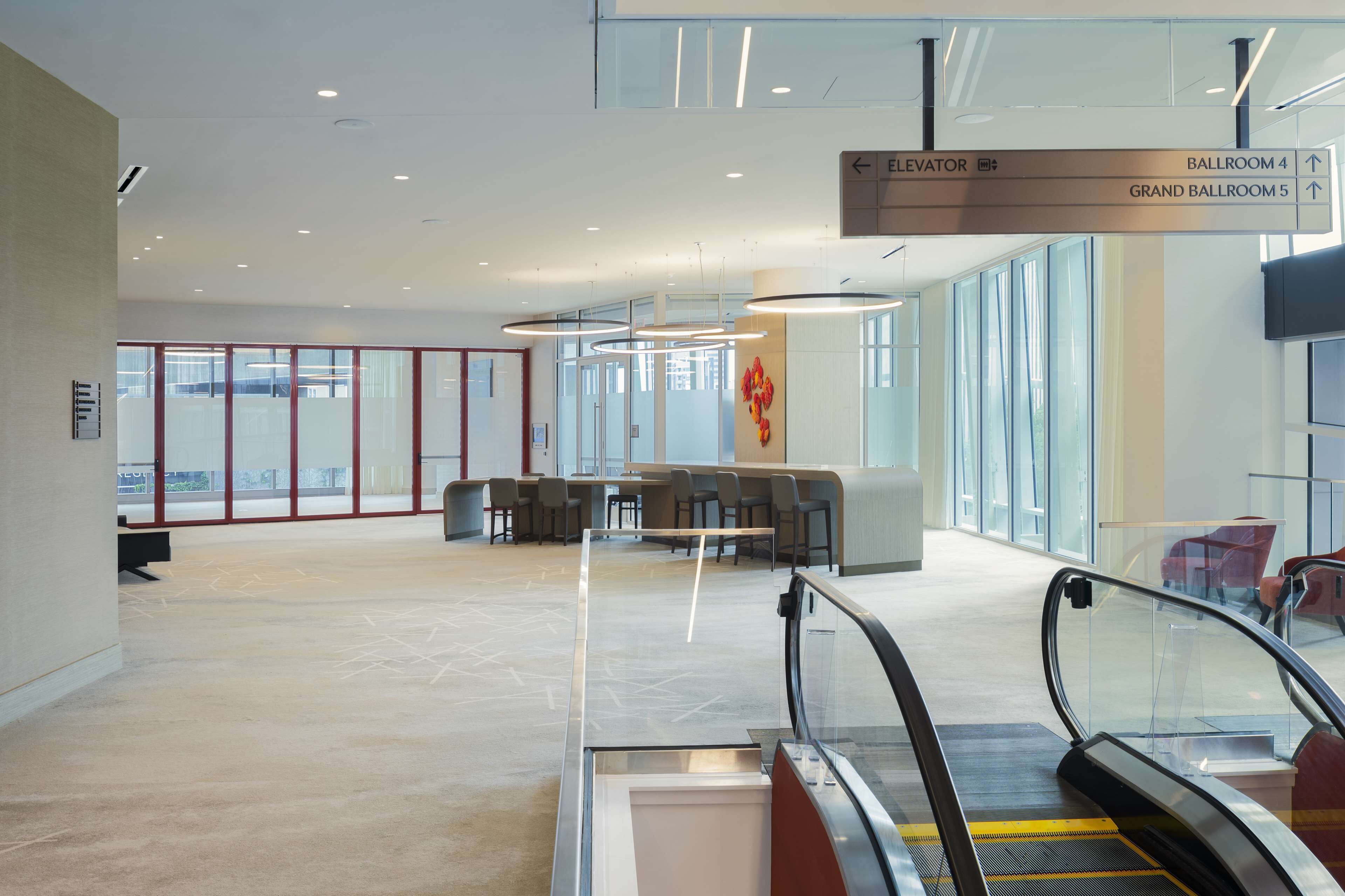 An interior view of a hotel lobby with an escalator, seating area, and directional signage for the elevator and ballrooms.