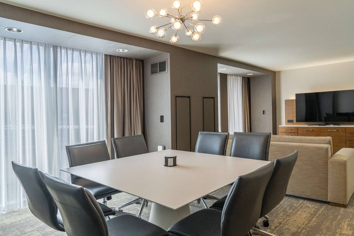 A modern hotel room with a large dining table, comfortable seating, a flat-screen TV, and a large window with curtains.