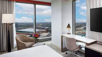 A hotel room with a large window, a bed, a desk, a chair, and a TV.