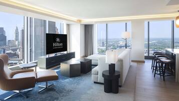 A modern hotel room with a large TV, comfortable seating, a dining area, and a city view.
