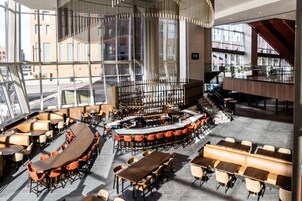 A modern restaurant with a central bar, multiple seating areas, and large windows offering views of the city.
