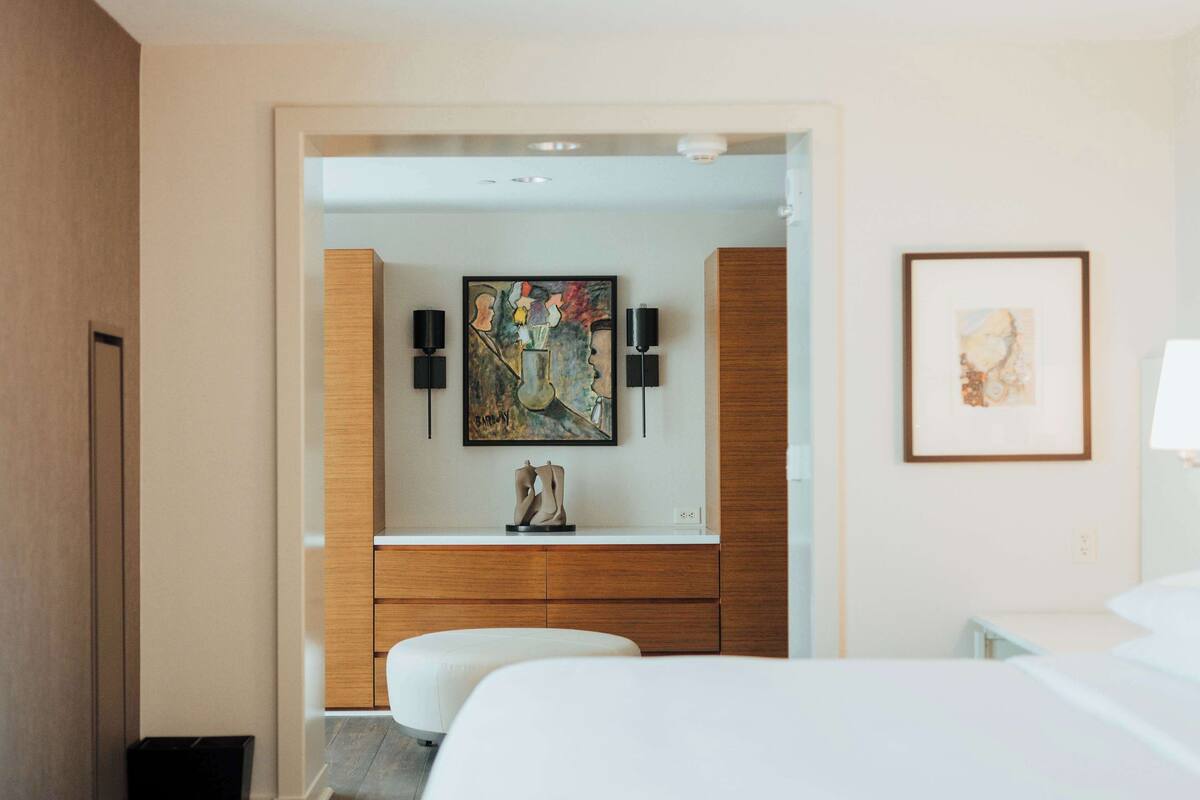 A hotel room with a wooden vanity, a painting above the mirror, and a bed with white bedding.