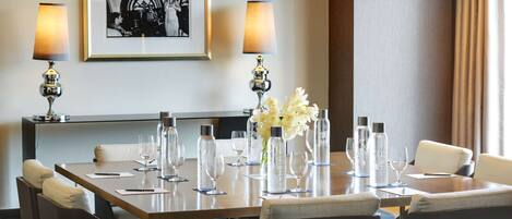 Een restaurantgedeelte van een hotel met een lange tafel gedekt voor een maaltijd, twee lampen, een ingelijst kunstwerk aan de muur en een bank op de voorgrond.