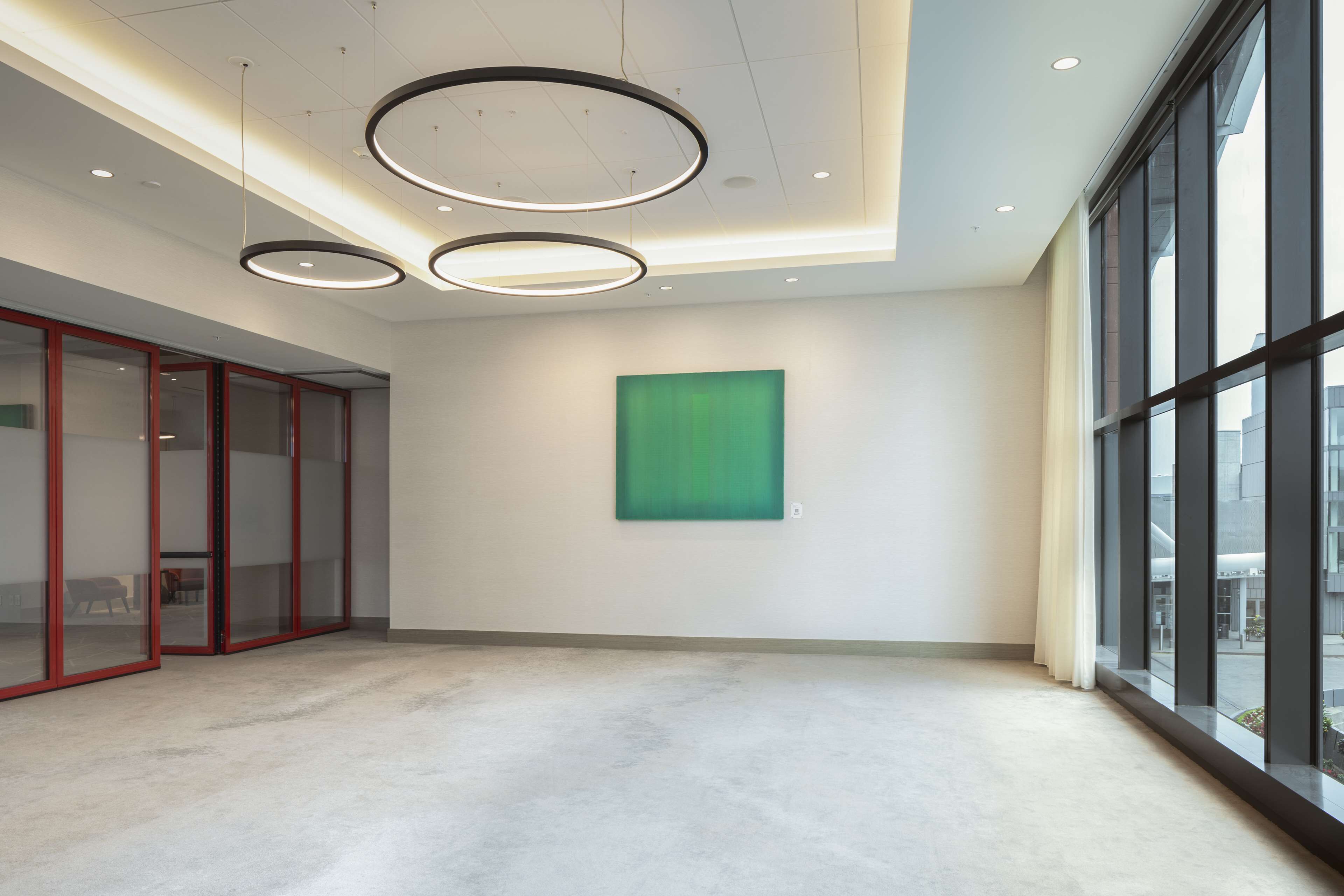 Een ruime kamer met een groot raam, een abstract schilderij in groene tinten en een moderne plafondlamp.
