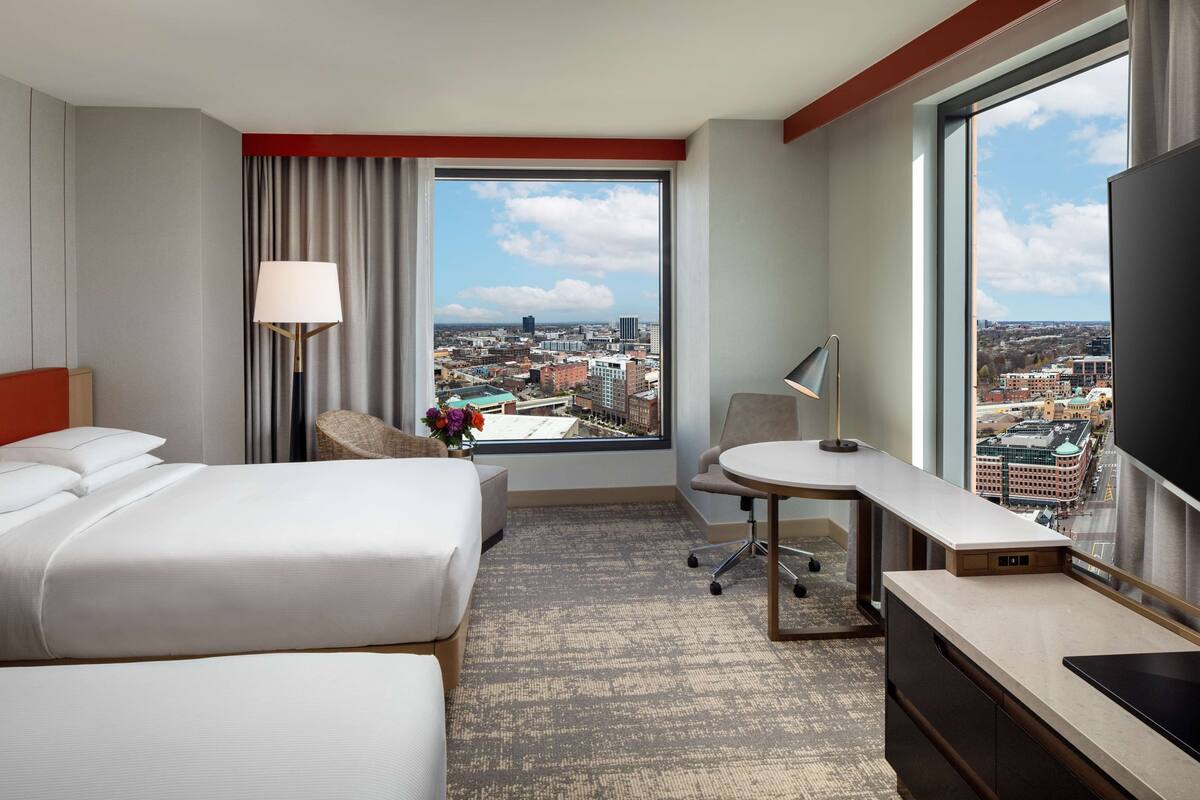 A hotel room with two beds, a desk, a chair, a lamp, and a large window offering a city view.