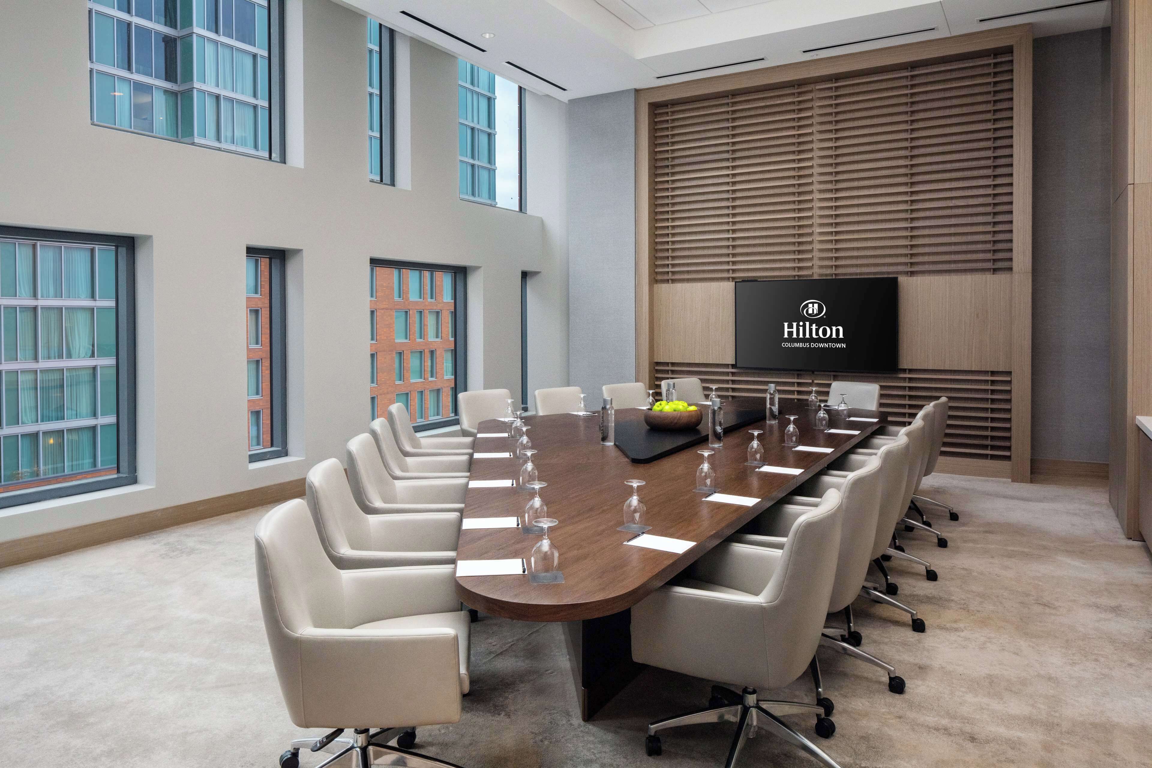 A conference room with a long table, white chairs, and a large screen displaying the Hilton Columbus Downtown logo.