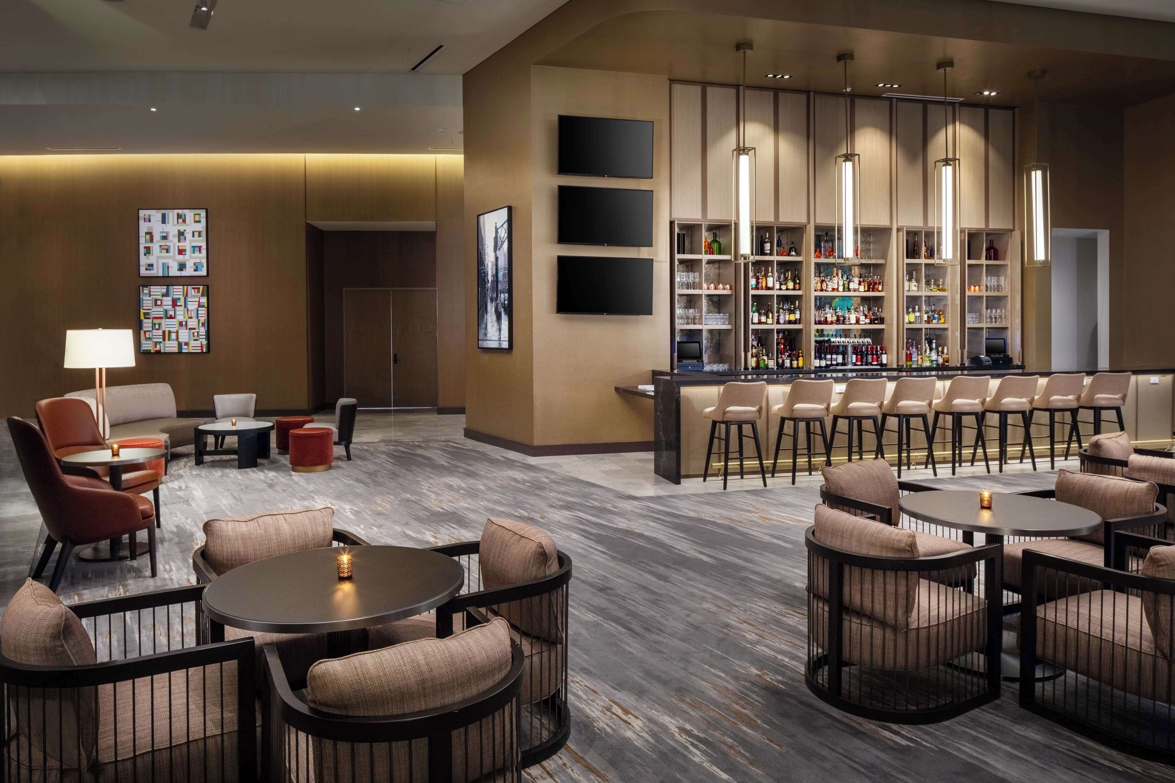 A modern lounge area with round tables and cushioned chairs, a bar with shelves of bottles, and a wall-mounted TV.