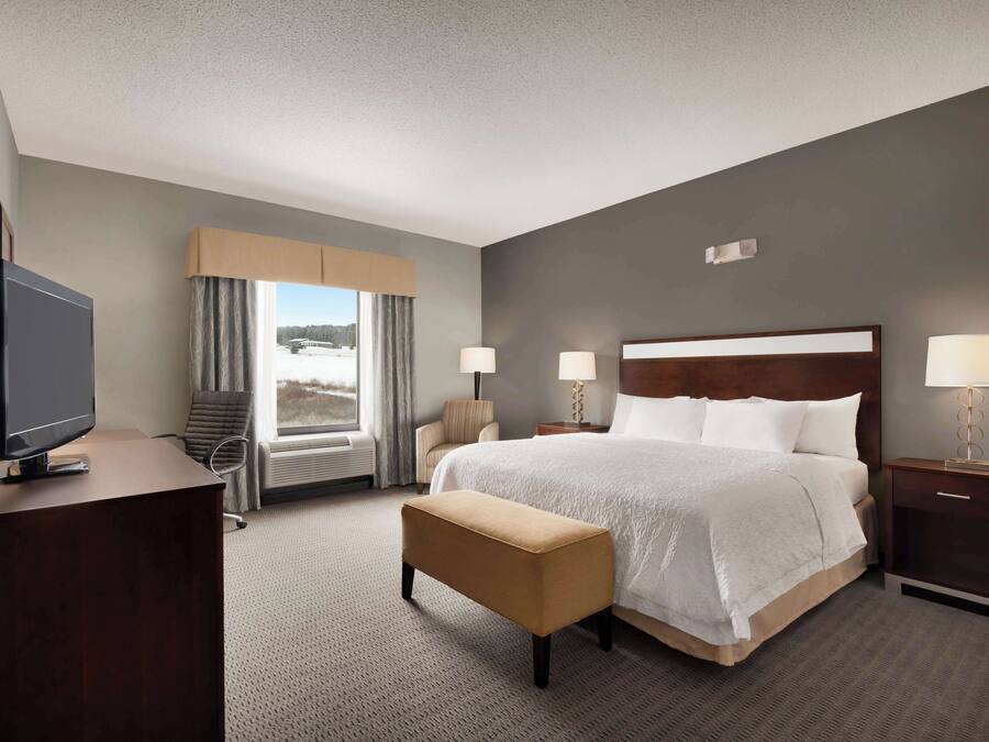 A hotel room with a large bed, a TV, a bench, and a view of a snowy landscape through the window.