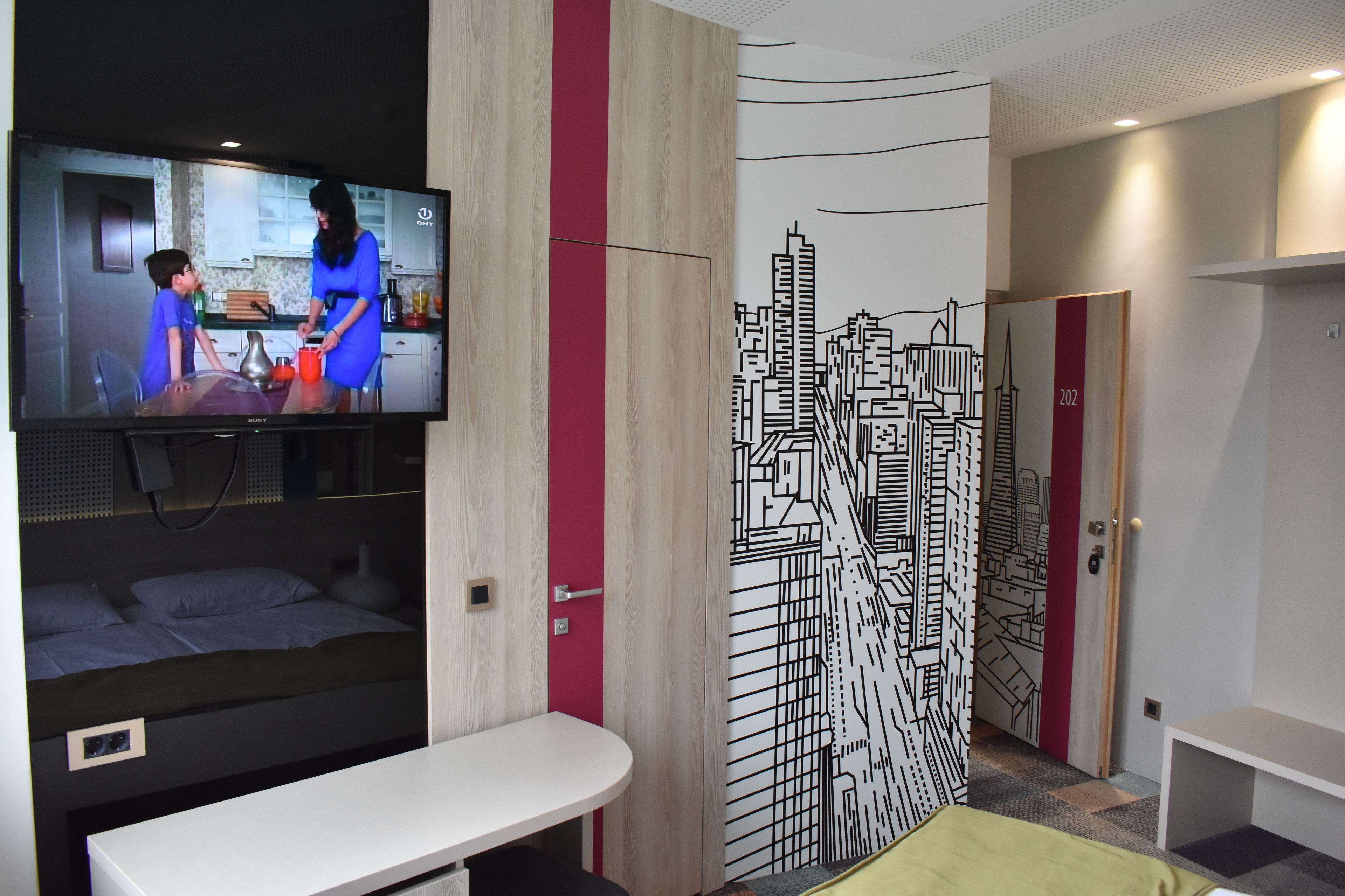 A modern hotel room with a TV, a bed, a desk, and a cityscape mural.