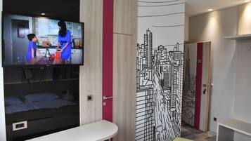 A modern hotel room with a TV, a bed, a desk, and a cityscape mural.