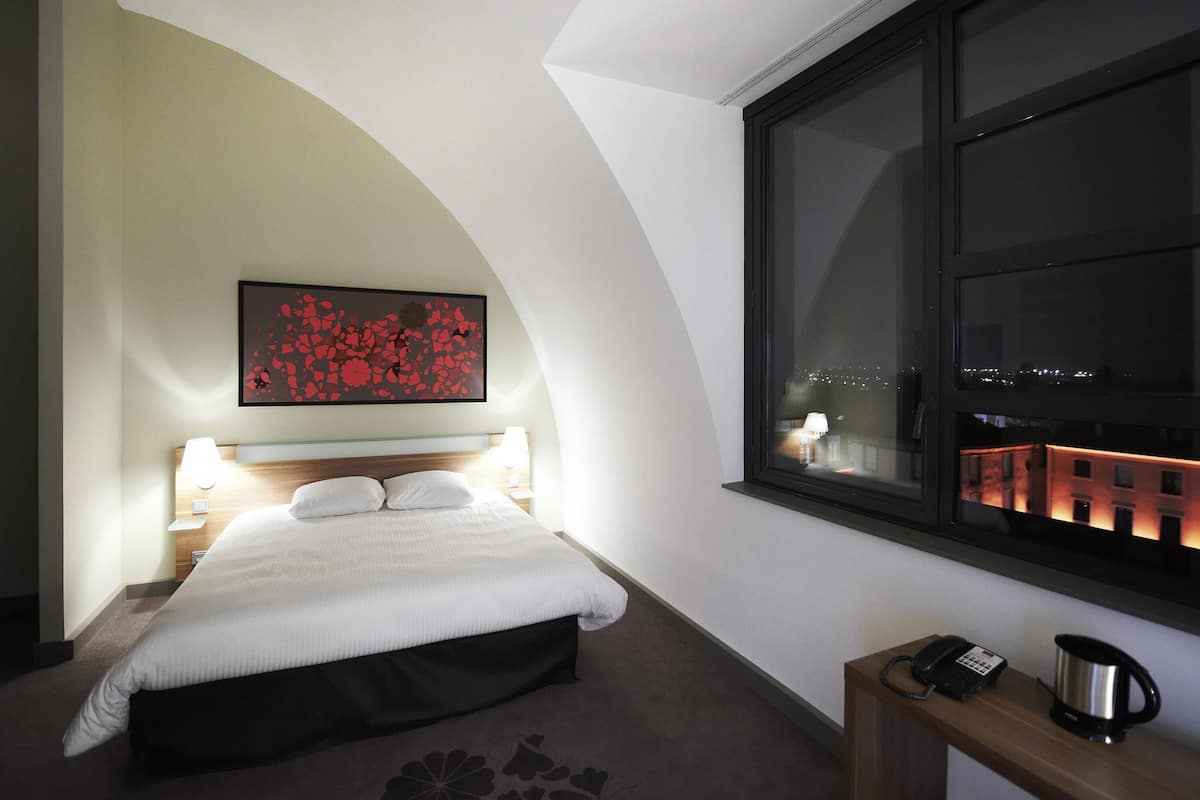 A hotel room with a bed, a nightstand with a lamp, a painting on the wall, a window with a view of the city, and a small table with a phone and a kettle.