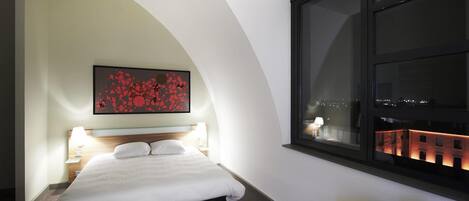 Une chambre d’hôtel avec un lit, une table de chevet avec une lampe, un tableau au mur, une fenêtre donnant sur la ville et une petite table avec un téléphone et une bouilloire.