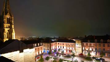 Vista nocturna de un edificio histórico con una torre de reloj, una plaza empedrada con árboles y edificios iluminados.