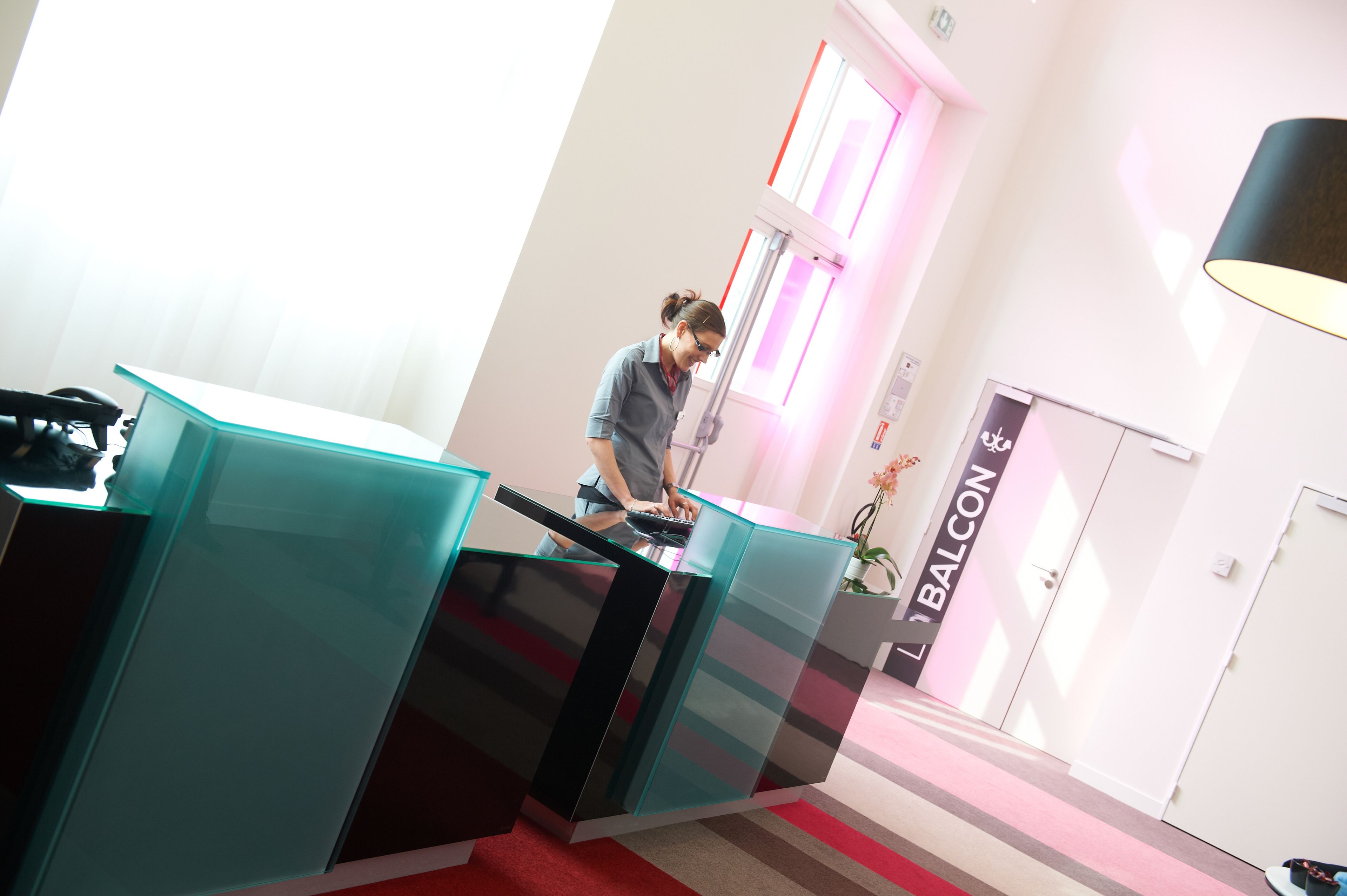 A reception desk with a modern design, a visible sign for 'BALCONY', and a large window with pink lighting.