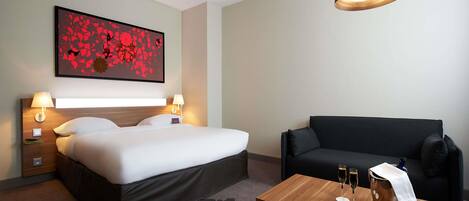 Ett hotellrum med en säng, en svart soffa, ett träbord med glas och en flaska, och en vägglampa.