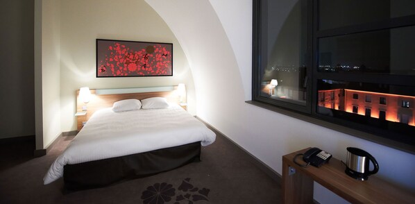 A hotel room with a bed, bedside lamps, a coffee maker, a telephone, and a window with a cityscape view.