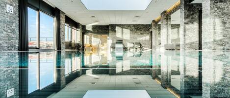 Una piscina interior moderna con paredes de vidrio, tragaluces y un muro de piedra.