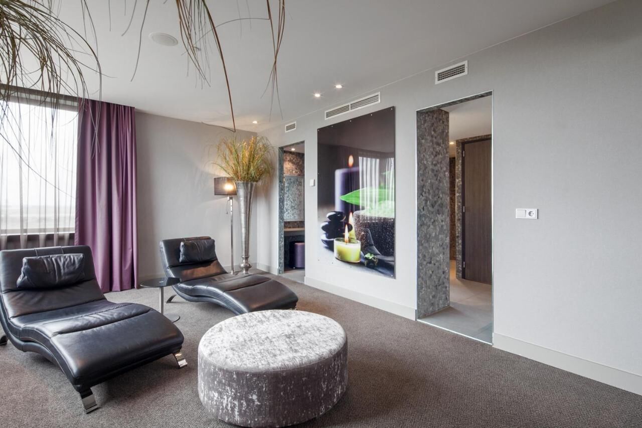 Un salon moderne avec un canapé, un pouf et une petite table. Il y a une grande fenêtre avec des rideaux, une plante et un téléviseur fixé au mur.