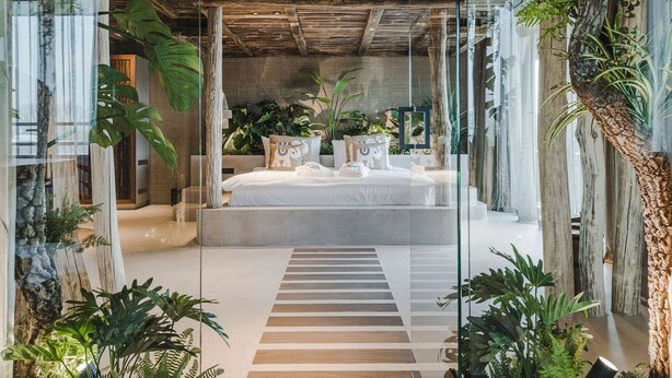 Een moderne slaapkamer met een groot bed, houten balken in het plafond en weelderige begroeiing.