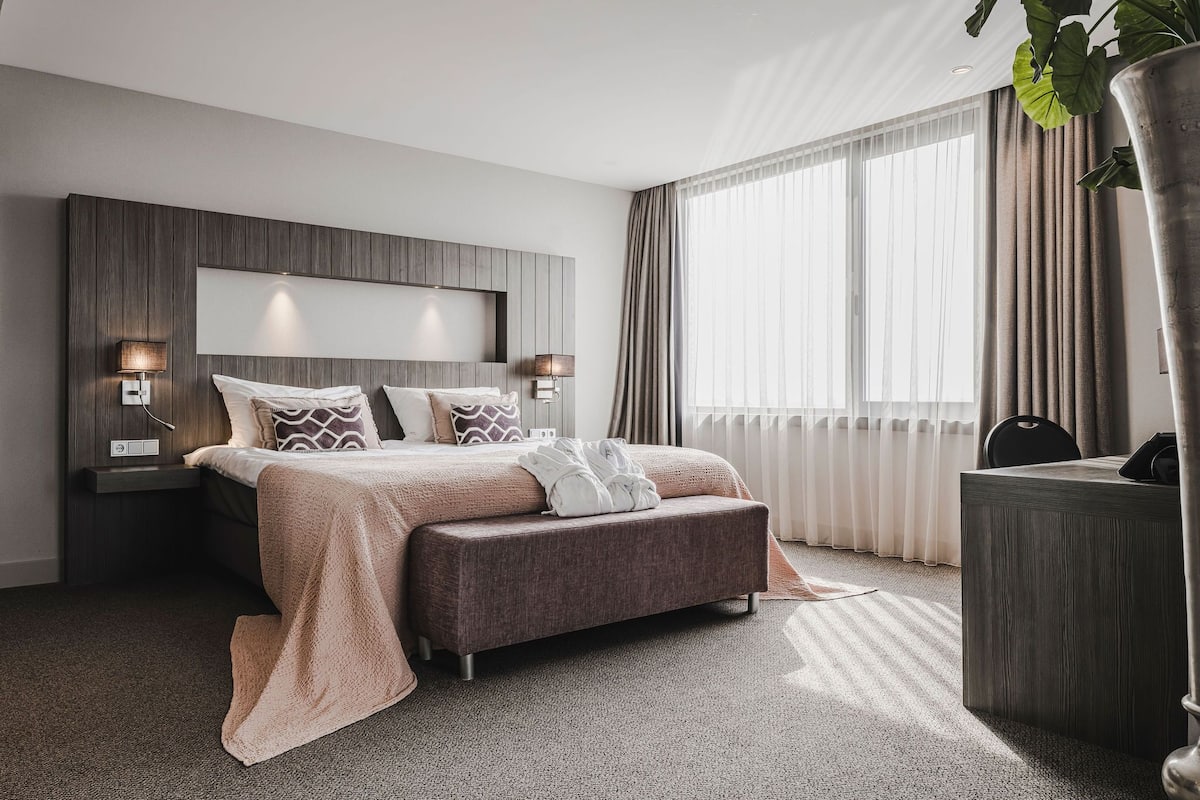 Moderni hotellihuone, jossa on suuri sänky, yöpöydällä olevat lamput, työpöytä ja ruukussa oleva kasvi.