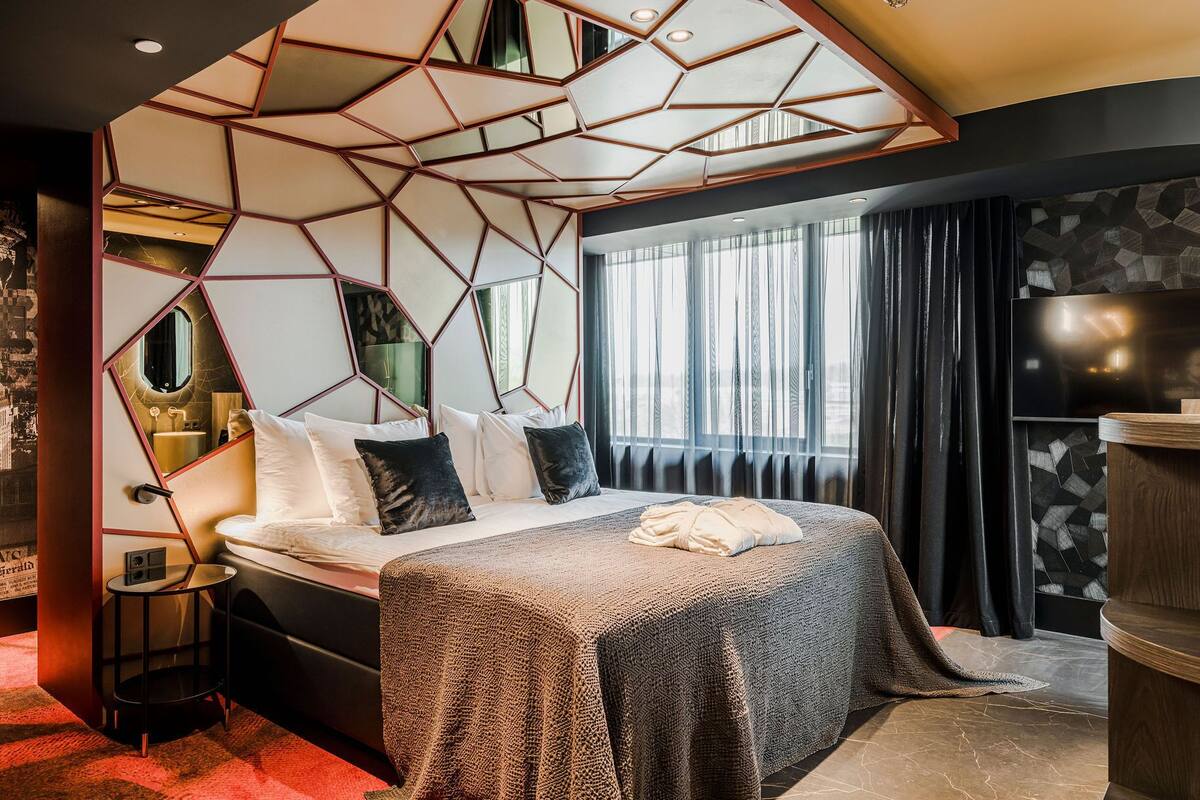Moderni hotellihuone, jossa on geometrisesti kuvioitu katto, suuri sänky, jossa on useita tyynyjä, pieni yöpöytä, televisio ja puinen sivupöytä.