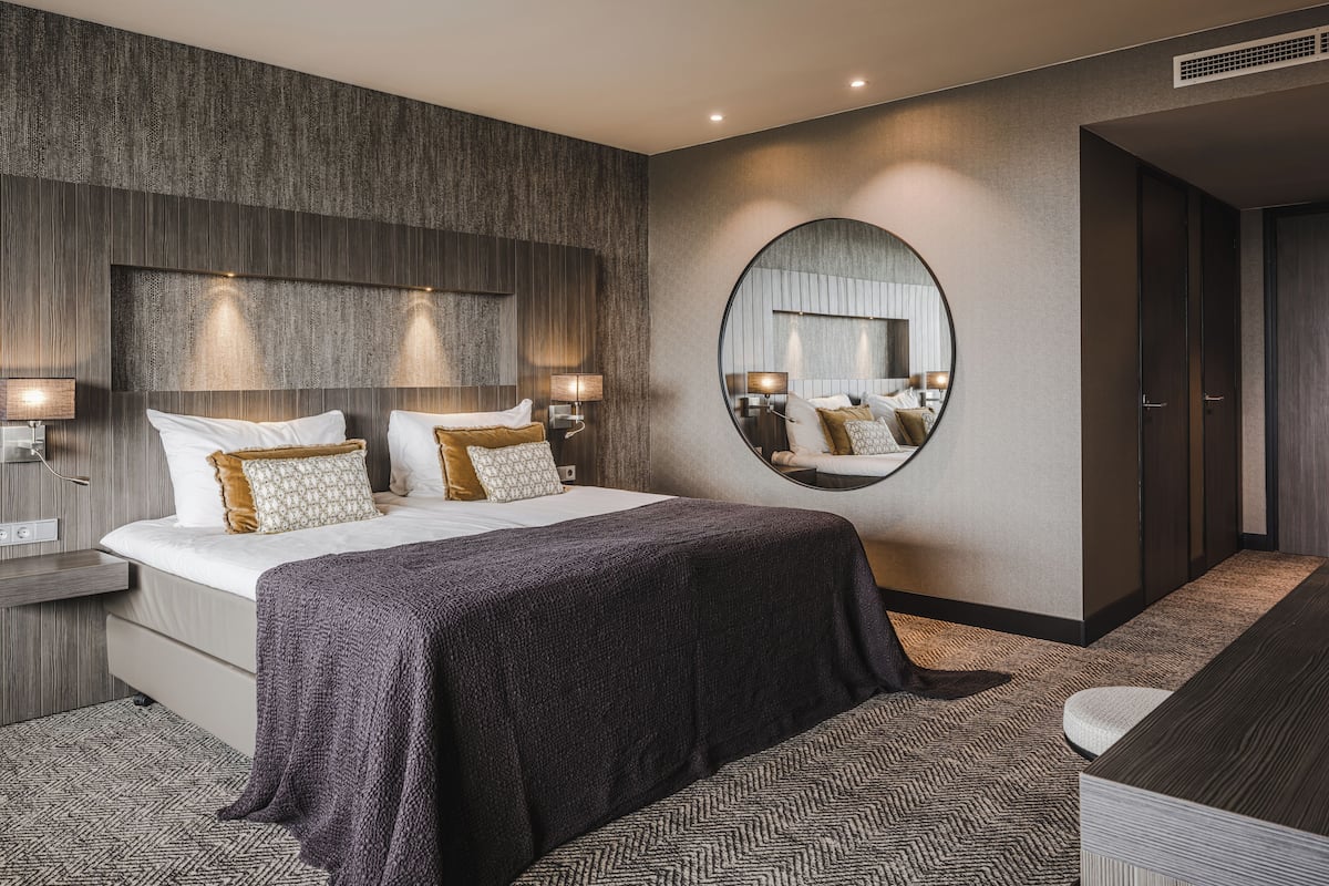 Moderni hotellihuone, jossa on suuri sänky, kaksi yöpöytää valaisimineen, pyöreä peili seinällä ja tummanvärinen matto.