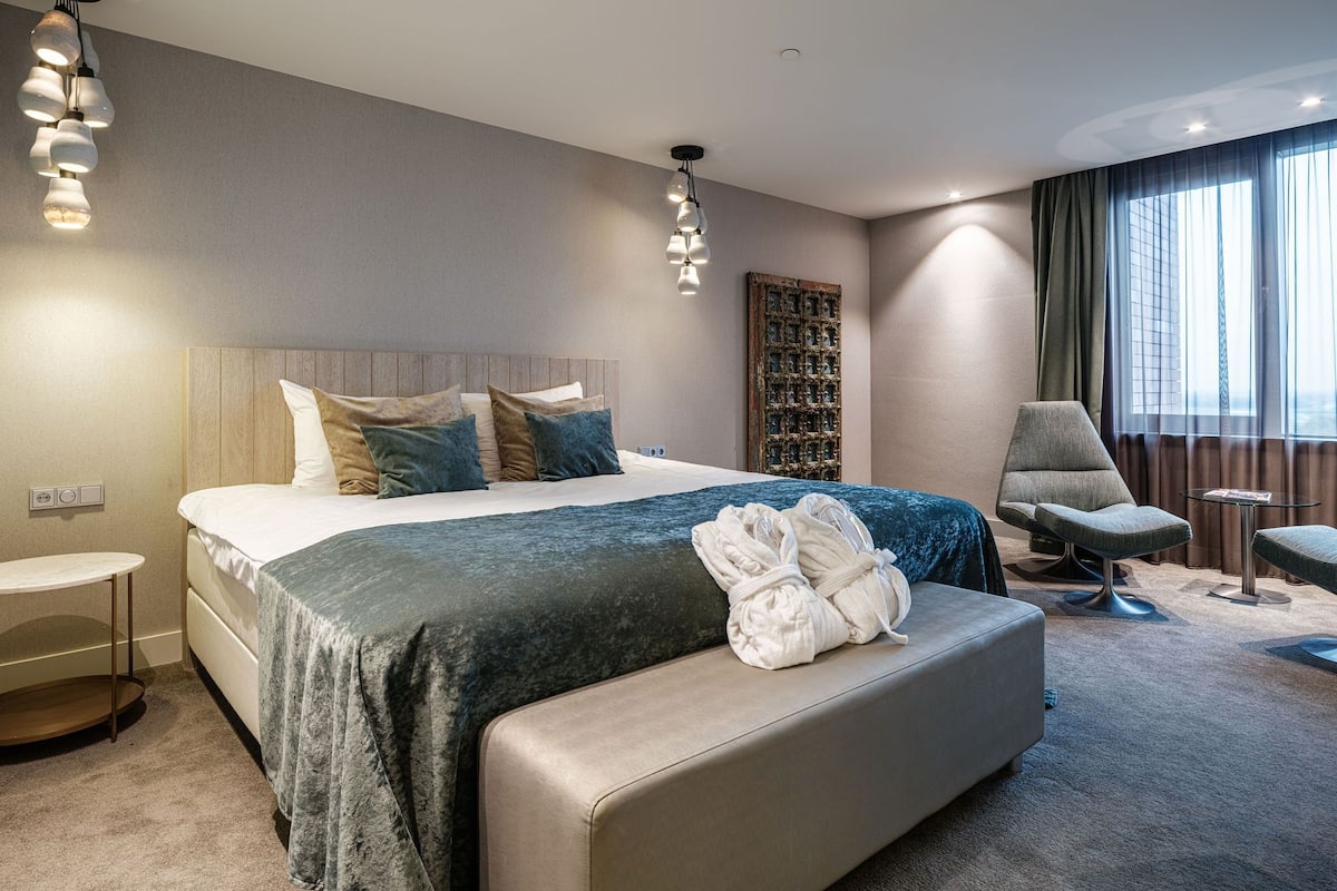 Moderni hotellihuone, jossa on suuri sänky, oleskelualue ja näkymä kaupunkiin.