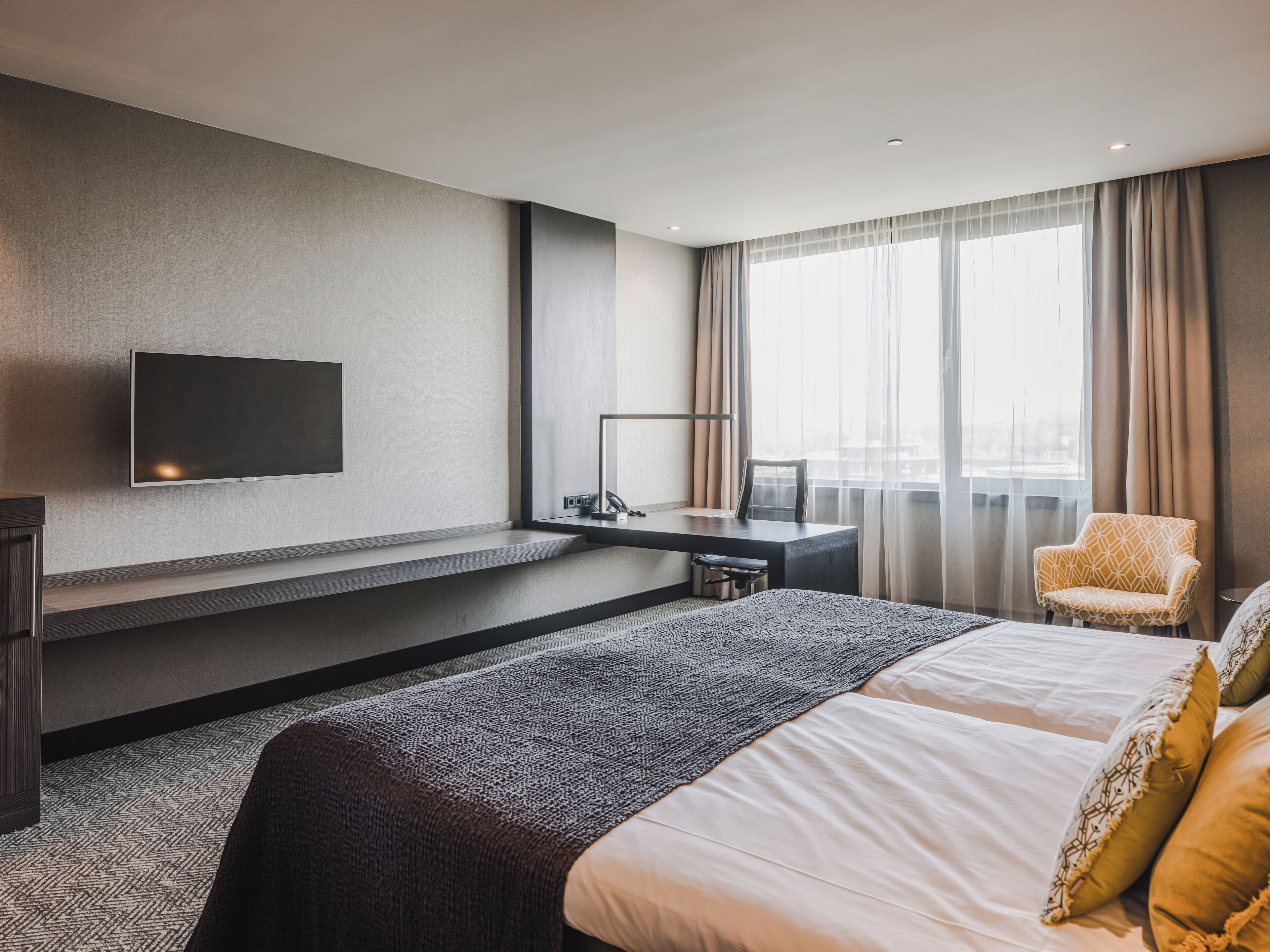 Quarto de hotel moderno com uma cama grande, uma escrivaninha, uma cadeira e uma televisão fixada na parede.
