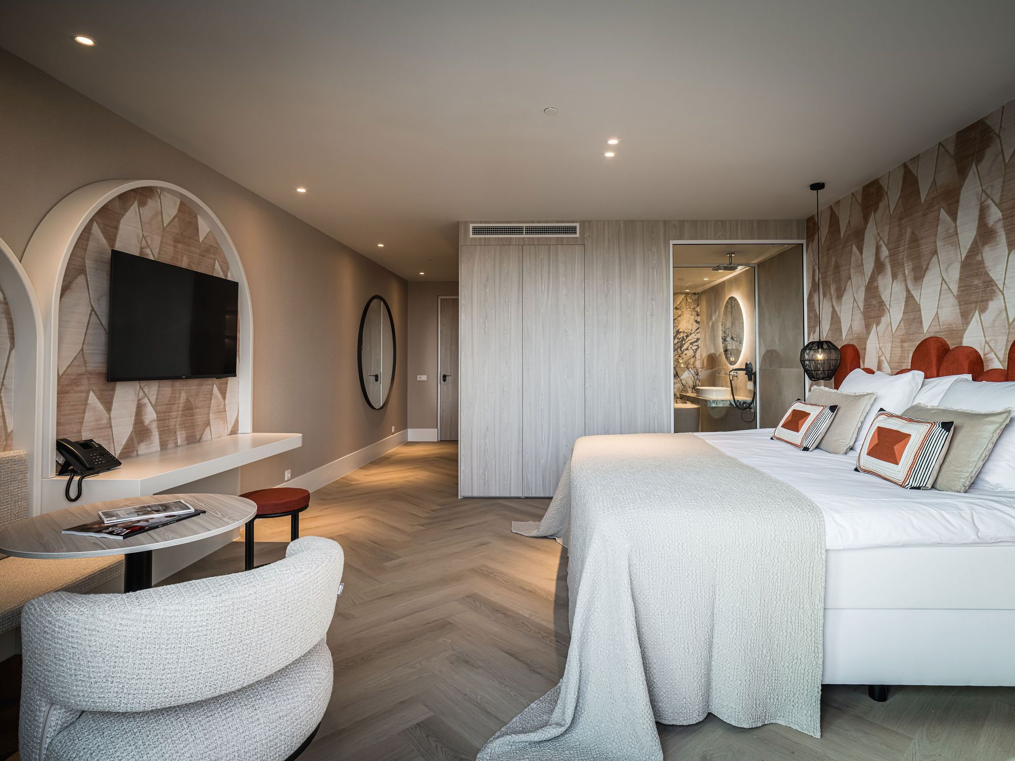 A modern hotel room with a large bed, a seating area with a table, a mounted TV, and a bathroom visible through an open door.