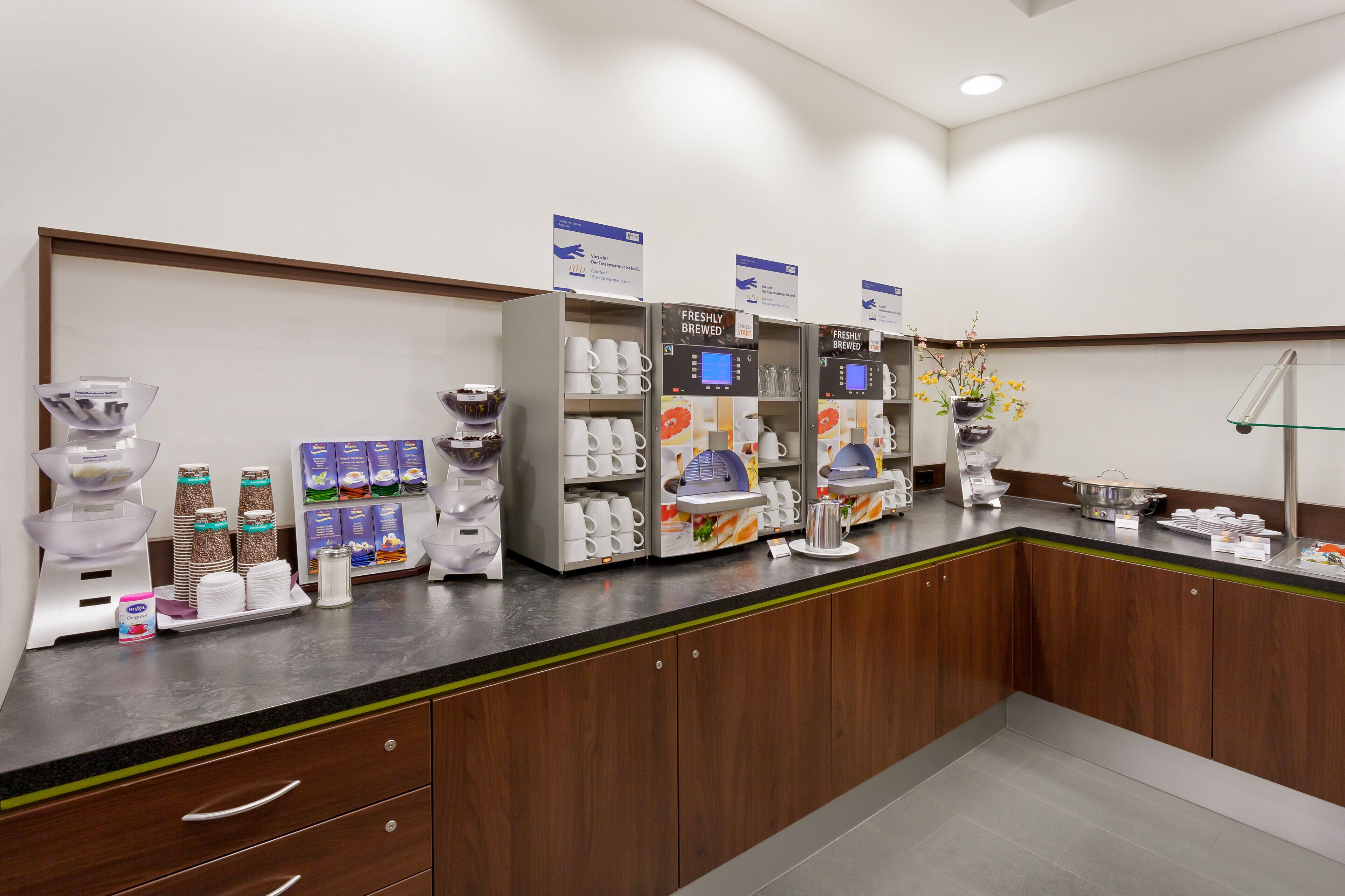 Een koffiehoek met meerdere koffiezetapparaten, een presentatie van koffiecups en een divers assortiment aan mokken en bekers.