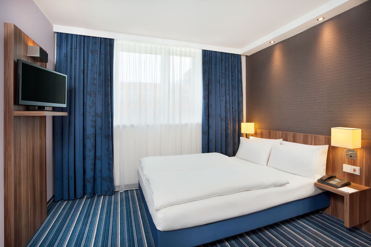 A hotel room with a large bed, a TV mounted on the wall, a bedside table with a lamp, and a window with sheer curtains.