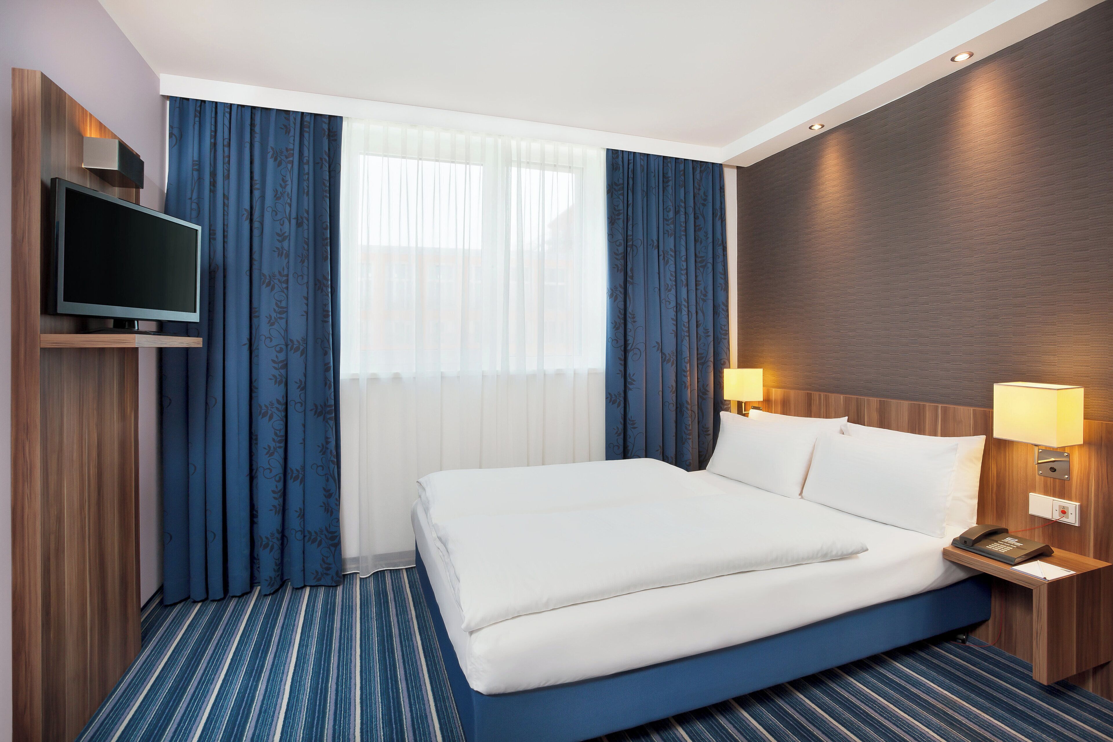 Una habitación de hotel con cama, televisor, mesita de noche con lámpara y ventana con cortinas.