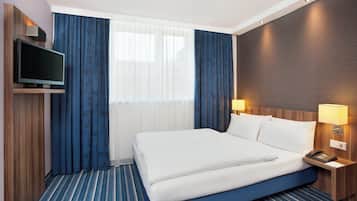 Quarto de hotel com cama, televisão, mesa de cabeceira com abajur e janela com cortinas.