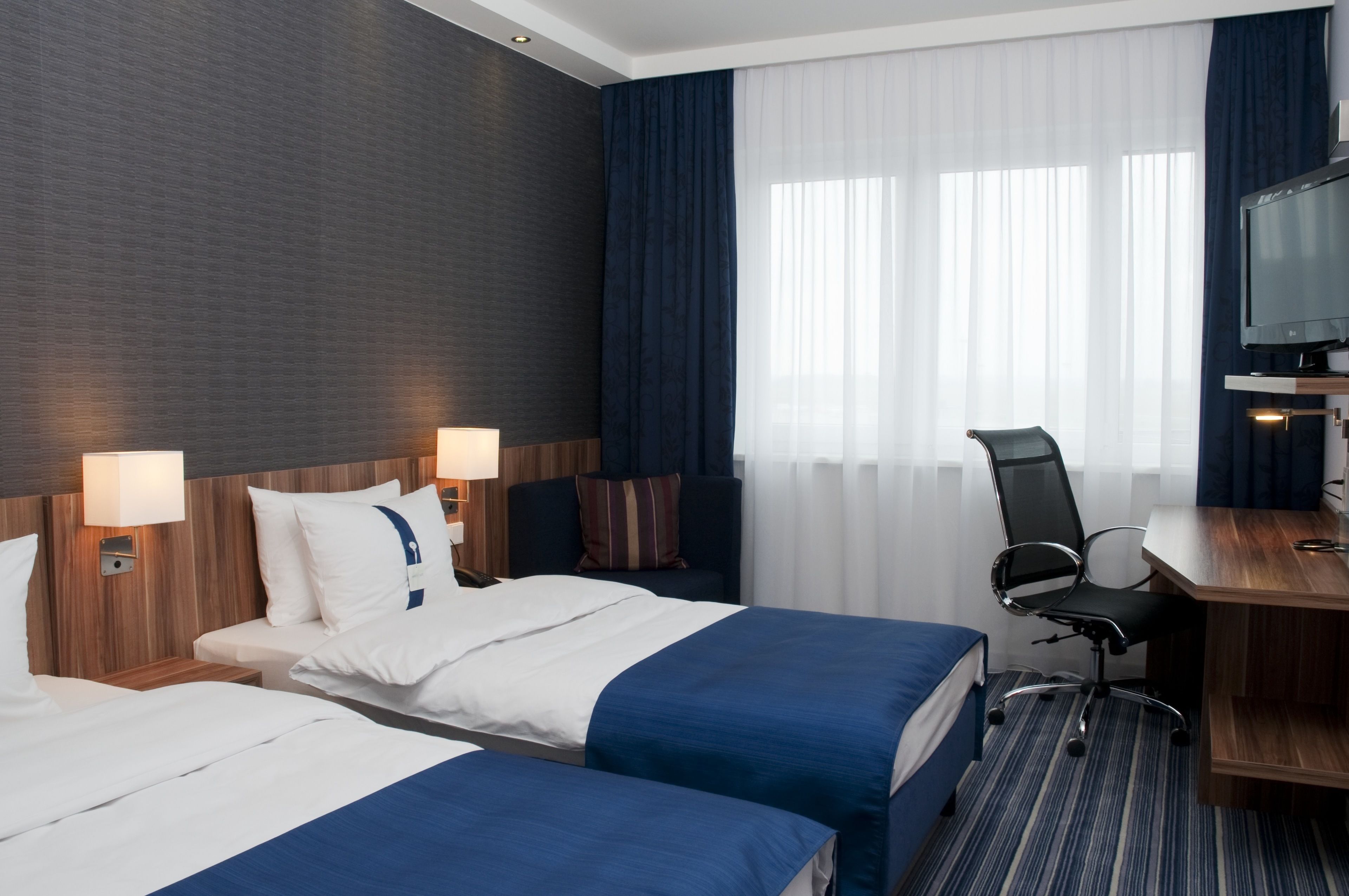 A hotel room with two beds, a desk, a chair, a TV, and a window with curtains.