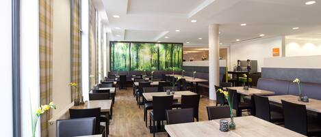 A modern restaurant with wooden tables and black chairs, large windows, and a forest mural on the wall.